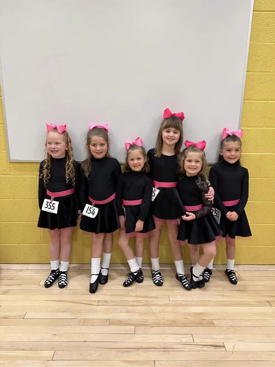 Six beginner dancers in black dresses and pink bows lined up before a competition