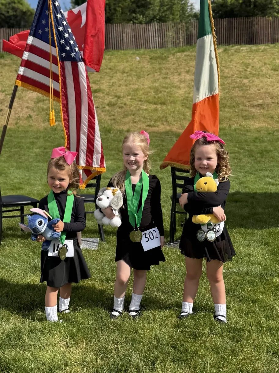 Three young dancers holding medals and stuffed animals in front of flags