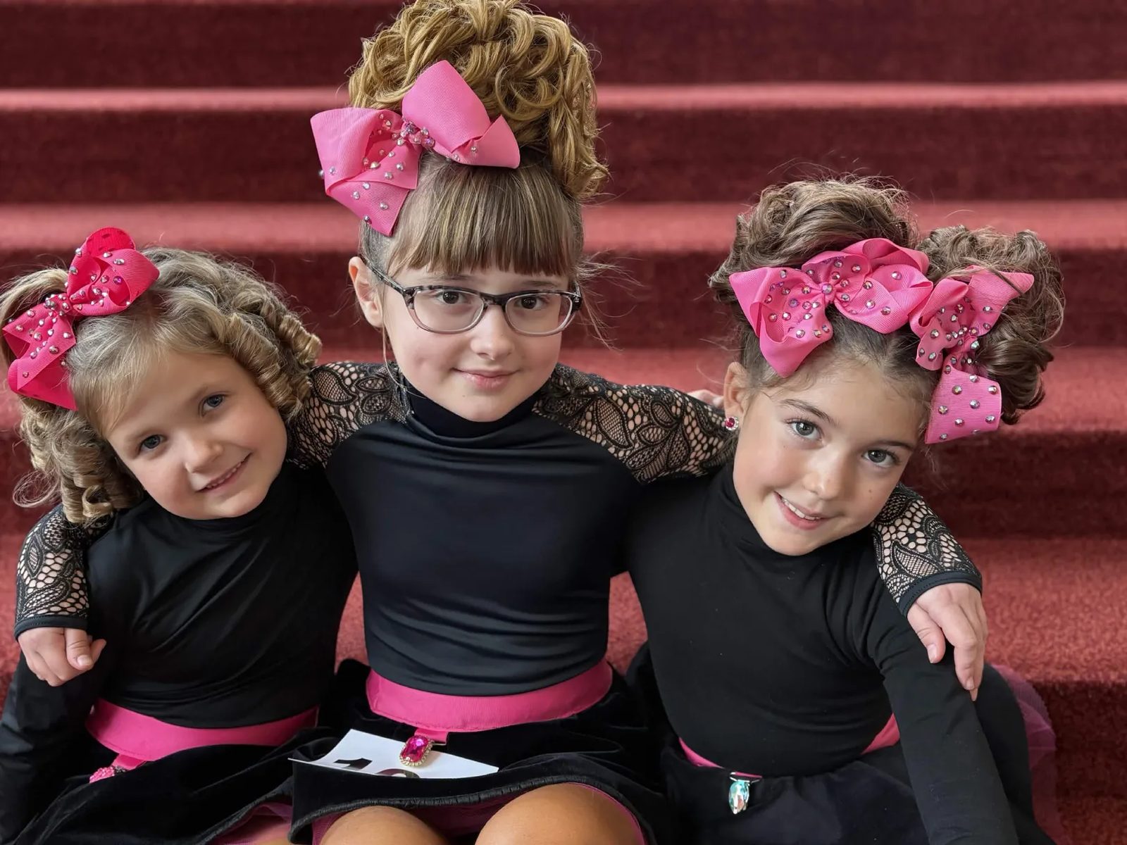 Three young Spirited Soles dancers with pink bows sitting together on red stairs at a competition